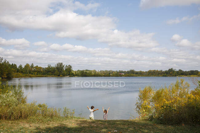 Vista distante de duas crianças felizes com braços levantados pelo lago — Fotografia de Stock