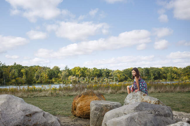 A small child in sundress sits on large rock by lake in summer — Stock Photo