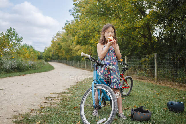 A little girl on bike takes break to eat apple in summer — Stock Photo