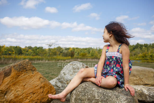 Small windblown girl with bare feet sits on boulder outside in summer — Stock Photo