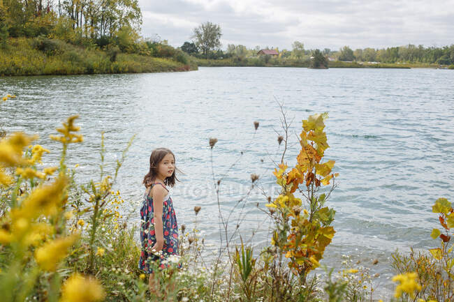 Petite fille regarde en arrière du bord du lac par champ de fleurs sauvages — Photo de stock