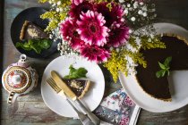Tarta de ganache de chocolate en el soporte de la torta y rebanada en el plato decorado con florero de flores y tetera vintage en la mesa - foto de stock