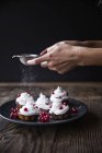 Immagine ritagliata di mani femminili spruzzando zucchero a velo su cupcake appena sfornati decorati con ribes rosso sul piatto — Foto stock