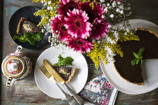Chocolate Ganache tart on cake stand and slice on plate decorated with vase of flowers and vintage teapot on table — Stock Photo