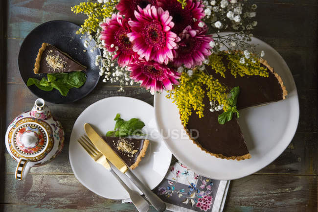 Chocolate Ganache tart on cake stand and slice on plate decorated with vase of flowers and vintage teapot on table — Stock Photo