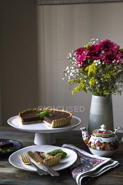 Tarte Ganache au chocolat sur support de gâteau et tranche sur assiette décorée avec vase de fleurs et théière vintage sur table — Photo de stock