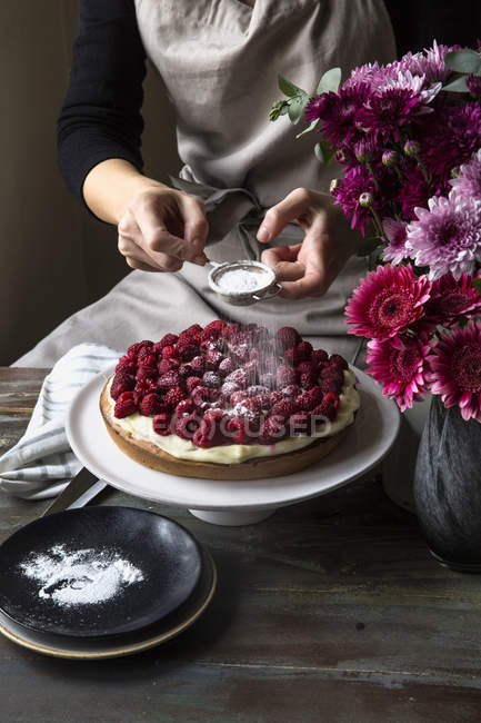 Partial view of female hands sprinkling icing sugar on cardamom cake with raspberries — Stock Photo