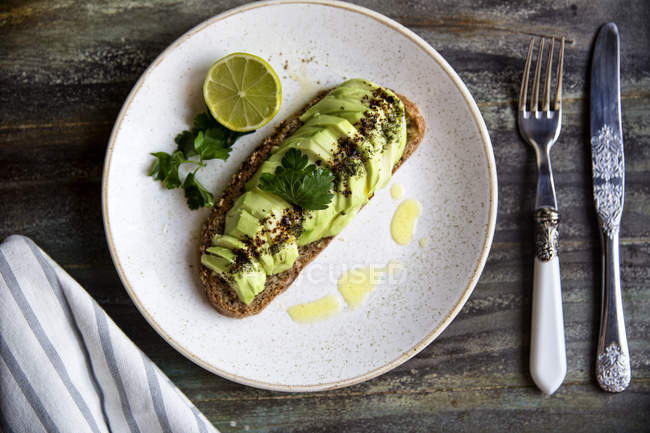 Plate of avocado toast with sauce, halved lime and herbs — Stock Photo
