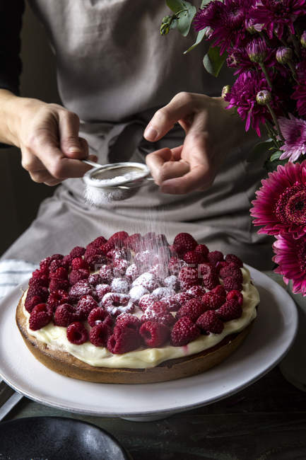 Vue partielle des mains féminines saupoudrer de sucre glace sur le gâteau à la cardamome avec des framboises — Photo de stock