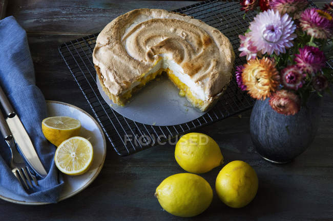 Lemon meringue pie on cooling rack with lemons and vase of flowers — Stock Photo