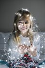 Glückliches kleines blondes Mädchen spielt und Weihnachtsbeleuchtung trägt, während es auf dem Bett sitzt — Stockfoto