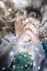 Little blonde girl playing and covering face with christmas lights while lying on bed — Stock Photo