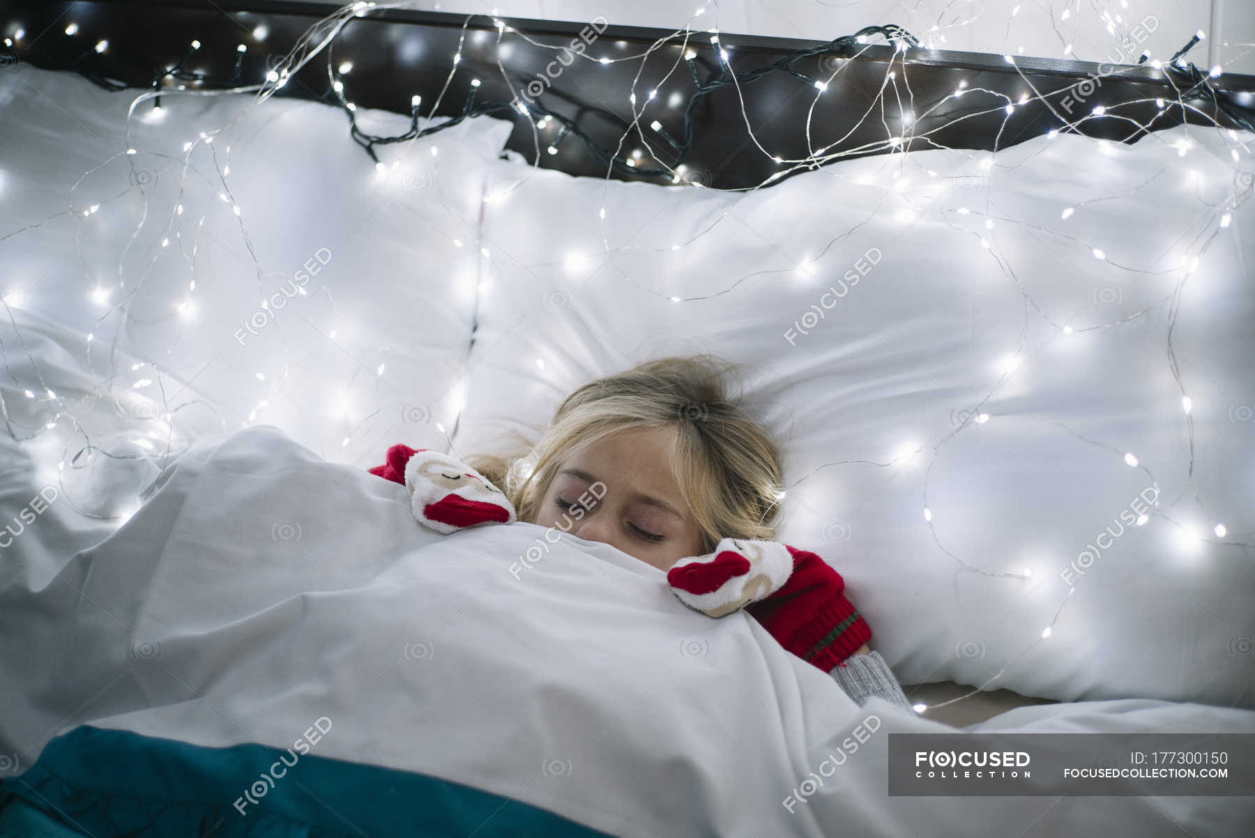 Little blonde girl lying in bed under the blanket, christmas garland with glowing light bulbs on