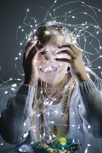 Happy little blonde girl playing and covering face with christmas lights — Stock Photo