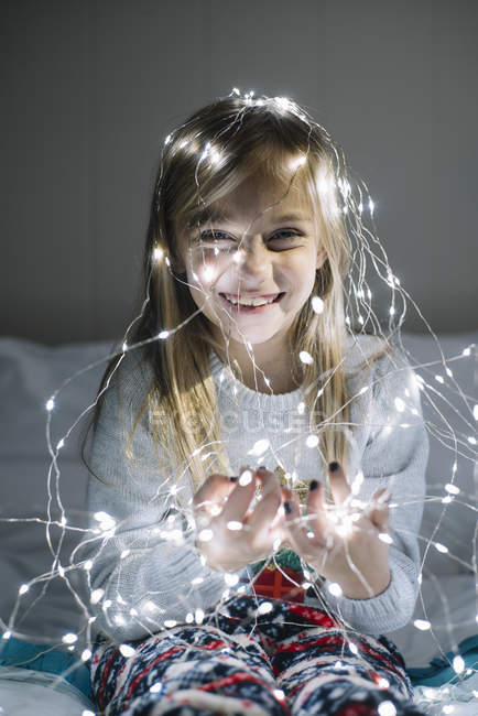 Feliz niña rubia jugando y usando luces de Navidad mientras está sentado en la cama - foto de stock