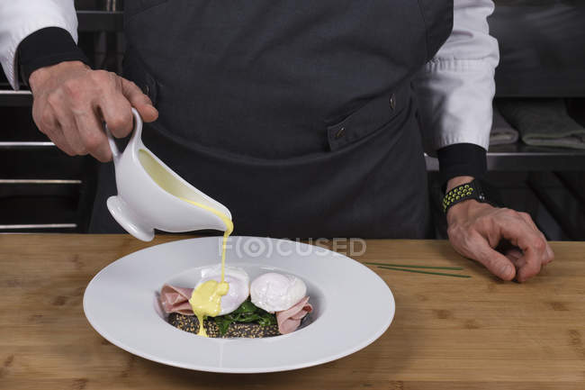 Vista cortada do chef masculino derramando molho em ovos benedict — Fotografia de Stock