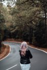 Vista posteriore di bionda giovane donna che cammina su strada vuota nella foresta — Foto stock