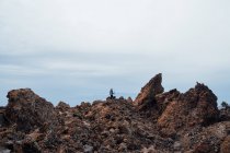 Далекий вид туриста, стоящего с селфи-палкой на скалистой местности под облачным небом — стоковое фото