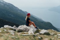 Vista laterale della donna bionda seduta sul masso sullo sfondo del pendio della montagna e del cielo — Foto stock