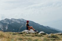 Side view of blonde woman sitting on boulder on background of mountain slope and cloudscape — Stock Photo
