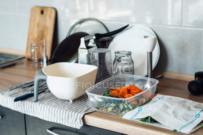 Conteneur avec légumes tranchés et ustensiles de cuisine sur comptoir de cuisine en bois — Photo de stock