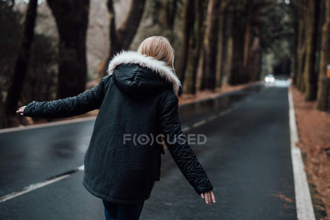 Vista posteriore di bionda giovane donna che cammina su strada vuota nella foresta — Foto stock