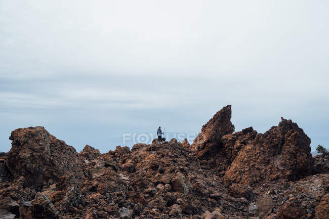Далекий вид туриста, стоящего с селфи-палкой на скалистой местности под облачным небом — стоковое фото