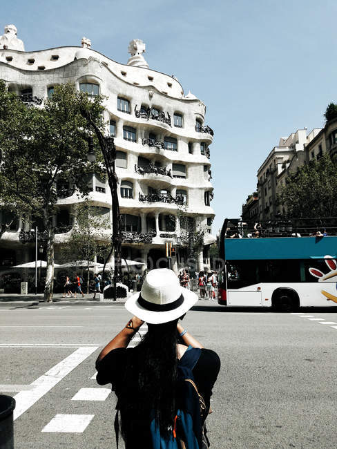 Vista trasera de la persona en sombrero blanco en la cruz peatonal tomando plano de la fachada del edificio - foto de stock