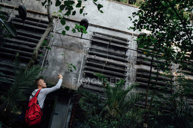 Vue latérale du jeune homme avec sac à dos prenant des photos de feuilles vertes — Photo de stock