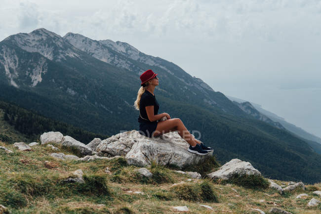 Side view of blonde woman sitting on boulder on background of mountain slope and cloudy sky — Stock Photo