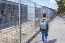 Rear view of schoolboy with backpack walking near fence at urban street — Stock Photo