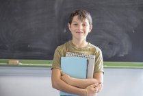 Porträt eines glücklichen Schülers, der mit Notizbüchern gegen die Tafel im Klassenzimmer steht — Stockfoto