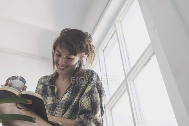 Mujer joven con camisa a cuadros sosteniendo libro y sonriendo - foto de stock