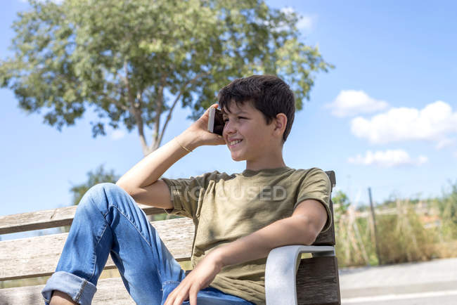 Schoolboy sentado no banco e falando no celular durante o intervalo — Fotografia de Stock