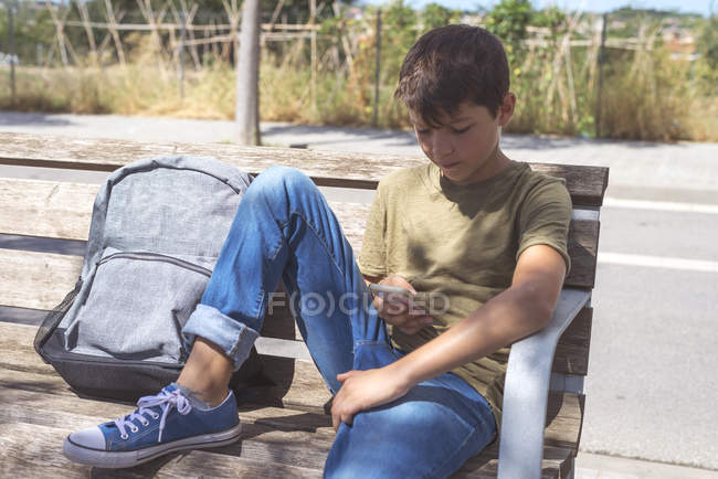 Écolier assis sur le banc et utilisant le téléphone mobile pendant la pause — Photo de stock
