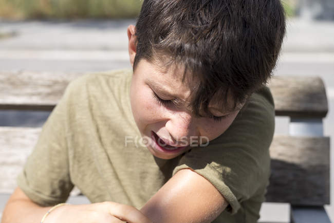 Portrait d'un garçon en pleurs avec une blessure à la main assis sur un banc dans un parc — Photo de stock