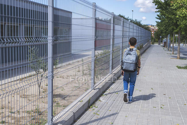 Вид сзади на школьника с рюкзаком, идущего рядом с забором на городской улице — стоковое фото