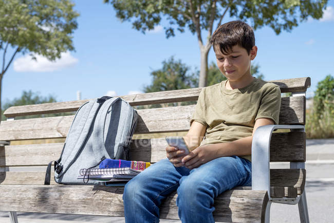 Schoolboy sitting on bench and using mobile phone during break — Stock Photo