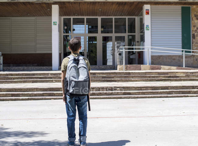 Вид сзади на школьного мальчика, стоящего и смотрящего на здание школы — стоковое фото