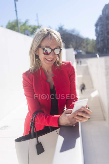Жінка в сонцезахисних окулярах використовує смартфон, спираючись на рейкові стіни — стокове фото