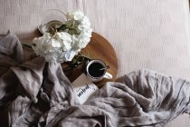 Enamel mug with coffee, white hydrangea flowers on wooden tray on bed — Stock Photo