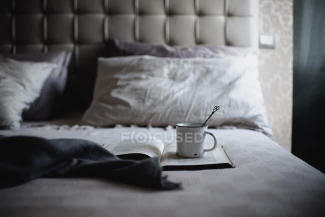 Эмалевая кружка с винтажной ложкой на открытой книге в постели — стоковое фото