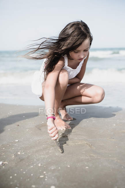 Chica agachándose en la playa y dibujando en la arena mojada . - foto de stock