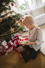 Pequeño chico rubio revisando regalos de Navidad sentado en el suelo - foto de stock
