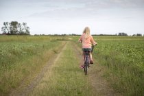 Loira menina andar de bicicleta ao longo da estrada de terra no campo verde — Fotografia de Stock