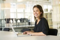 Молода жінка використовує ноутбук в офісі і посміхається — стокове фото