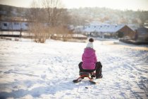 Visão traseira da menina trenó ao longo da estrada nevada com paisagem urbana no fundo — Fotografia de Stock