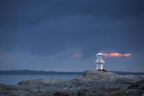 Illuminated lighthouse on rocks at dusk with cloudy sky — Stock Photo