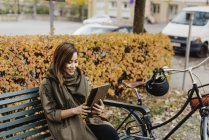Mujer joven sentada en el banco y usando la tableta digital cerca de la bicicleta - foto de stock