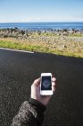 Hand holding smartphone with compass, road and seascape on background — Stock Photo